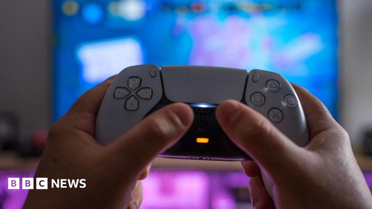 A white PlayStation 5 controller being held up by someone's hands against a blurred TV screen behind it