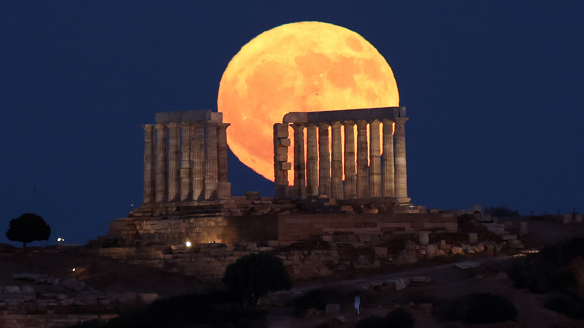 A large golden moon is seen over a series of ancient ruins