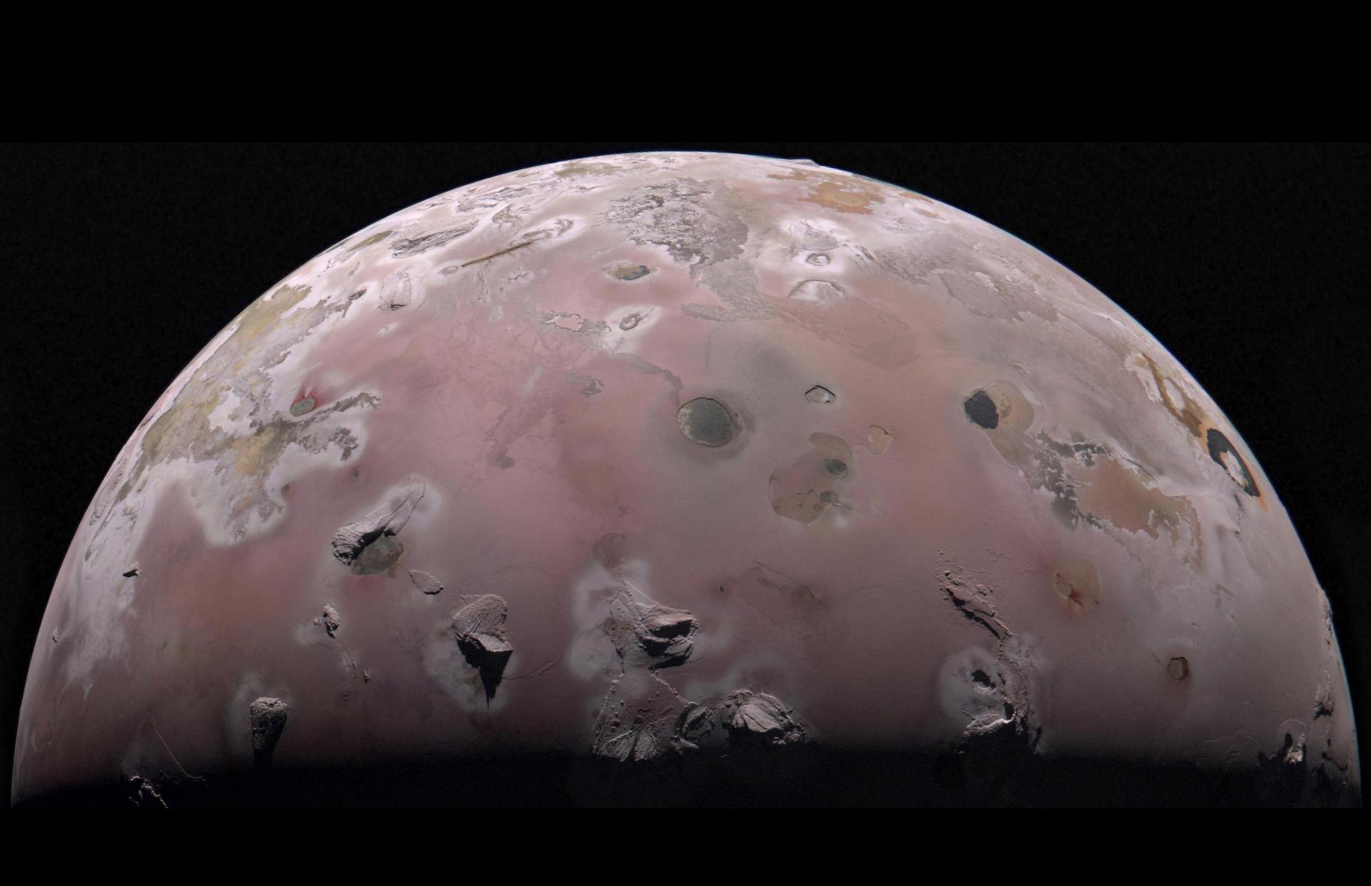 A view of half of the sphere of Jupiter’s volcanic moon Io against black space. The surface is colorful pinkish-tan and